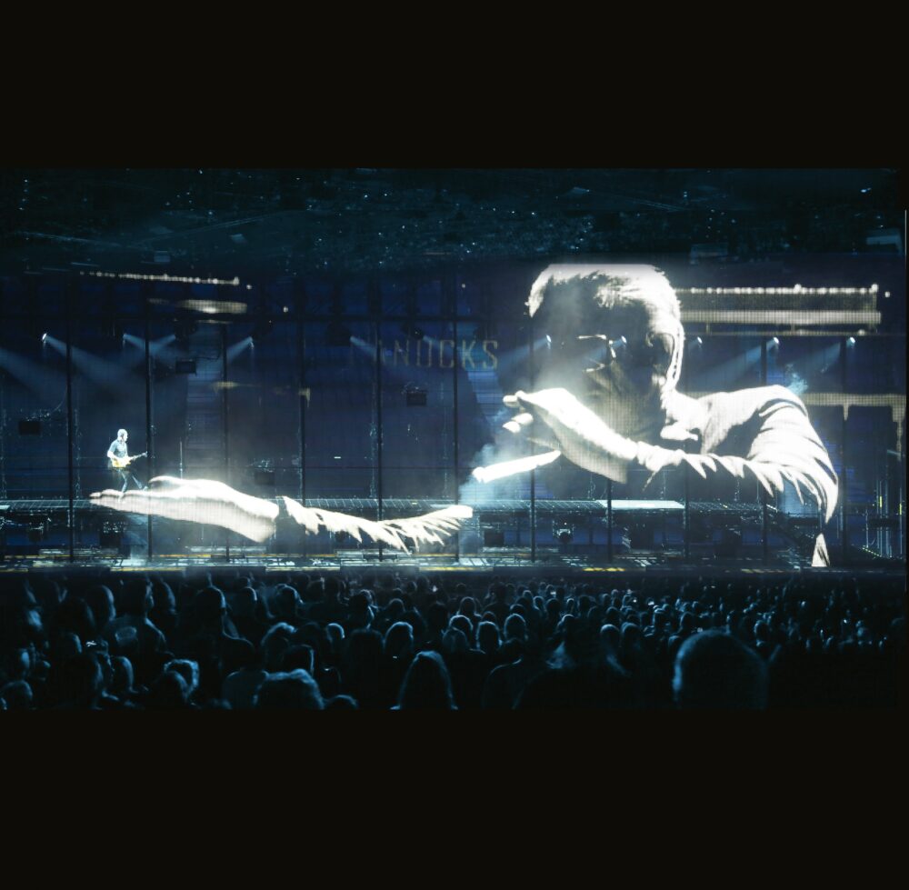 A crowd of people facing a screen with a zoomed-in image of a person singing and lifting up a much smaller person in their right hand.