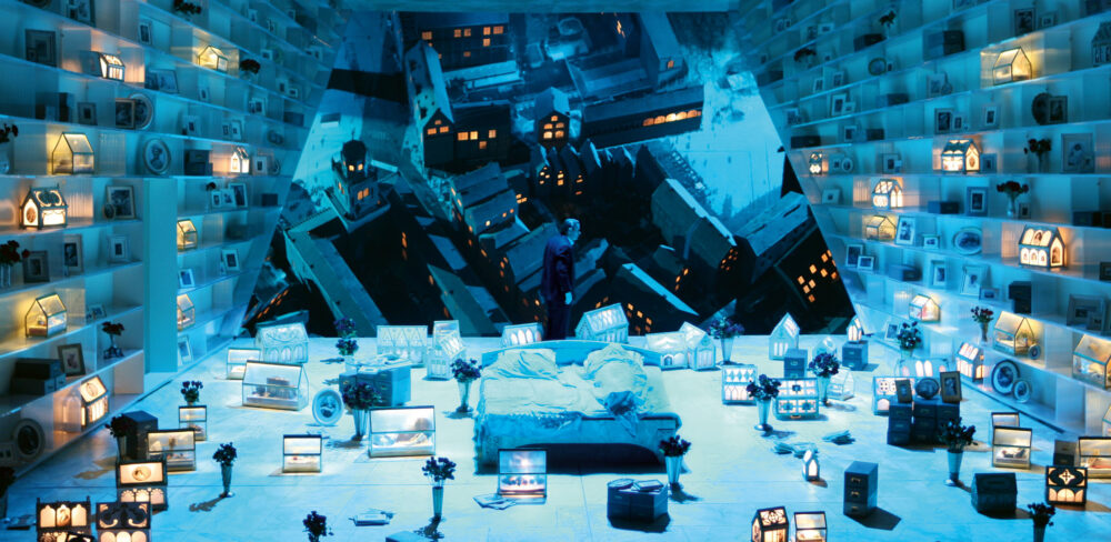 A person lying in a bed is at the center of a blue-lit room with slanted walls. Projected on the back wall is a dynamic image of floating buildings with yellow lights in the windows. The two other walls and floor are full of small house models with yellow lights, picture frames, and flowers in vases.