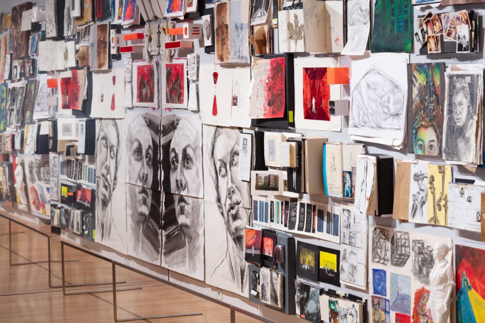 View from the right of a large-scale collage of artworks and sketchbooks pinned to a white wall. Some works are black and white whereas most are in color. Most of the imagery depicts figures, faces, and geometric forms.
