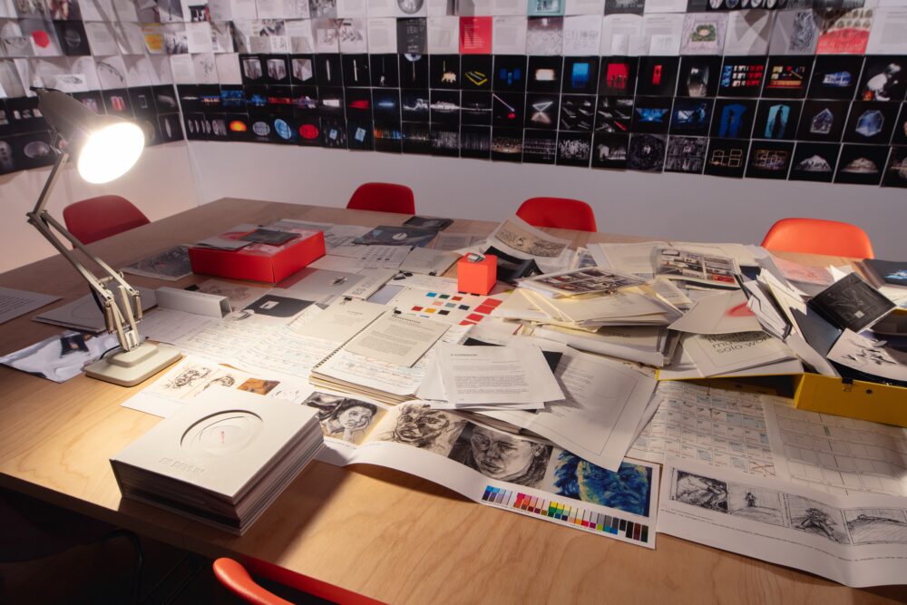 Two walls are shown in the background covered in square pages featuring text and images that are lined up next to each other. In the foreground is a large wooden table covered in more print materials, illuminated by a lamp; several red chairs surround the table.