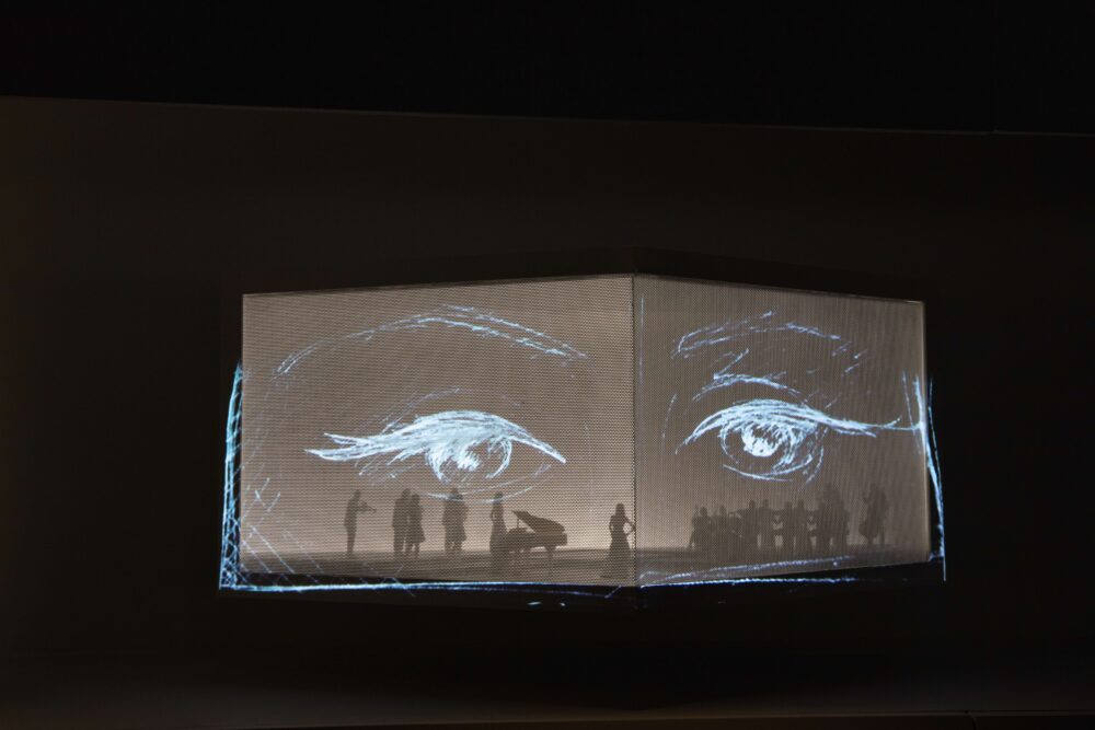 Slightly folded rectangular structure, above black background, with large-scale projection of a drawing of two opened eyes; inside the structure are faint small figures.