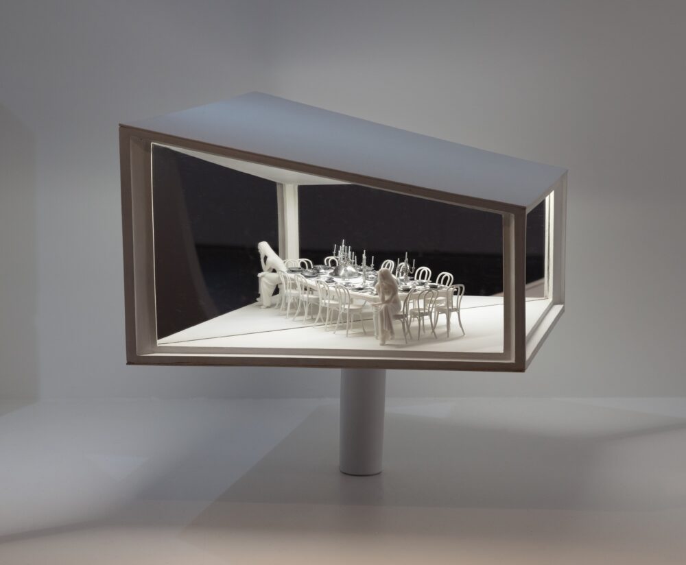 White model of a trapezoidal structure with a mirrored wall and white light glowing from inside, illuminating two figures sitting at opposite ends of a dining table.