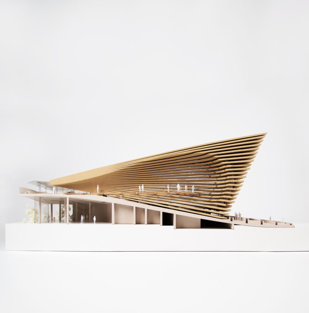 Model, made of wood and white materials, of an architectural structure seen from the side. A white rectangle is the base that holds the triangular structure made of horizontal lines. Small figures throughout the model indicate scale.