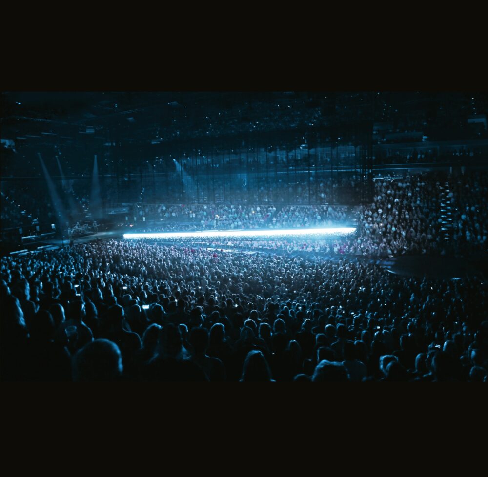 A large, indoor concert venue full of people with a long horizontal bar of glowing light floating above the crowd.
