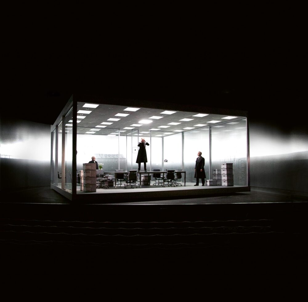Black rectangular structure with no walls housing office furniture and a ceiling with several squares of white light; inside are three people, one standing on the table. Behind the structure is a hazy stripe of white light that cuts across the black background.
