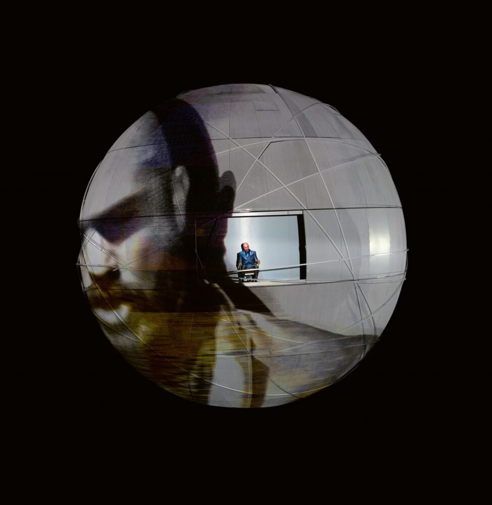 Spherical structure floating in front of a black background with a large-scale face projected on the surface. A person, dwarfed by the structure, stands within a rectangular cutout toward the middle of the structure.