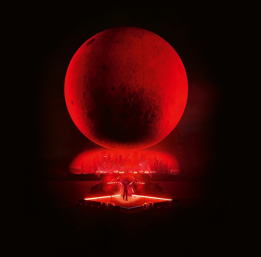A dark room with a large, red, moon-like sphere suspended above a glowing red stage with a relatively small person on it.