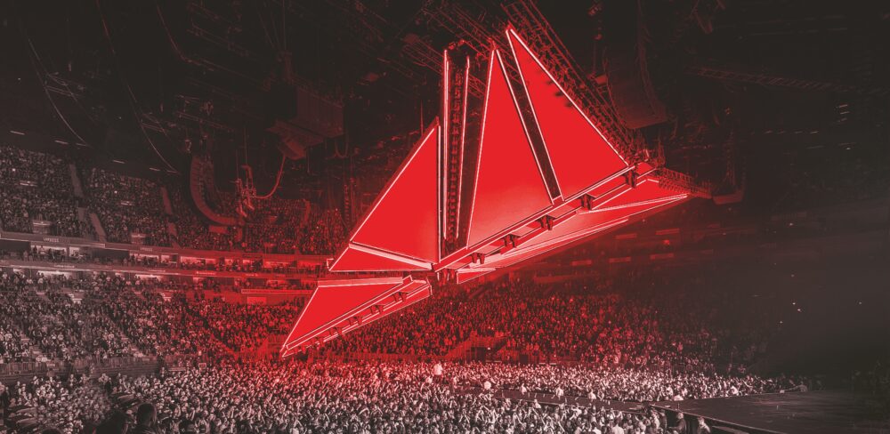 A large, indoor concert venue full of people with a series of large red triangles that together form a spaceship-like structure, suspended above the crowd at an angle.