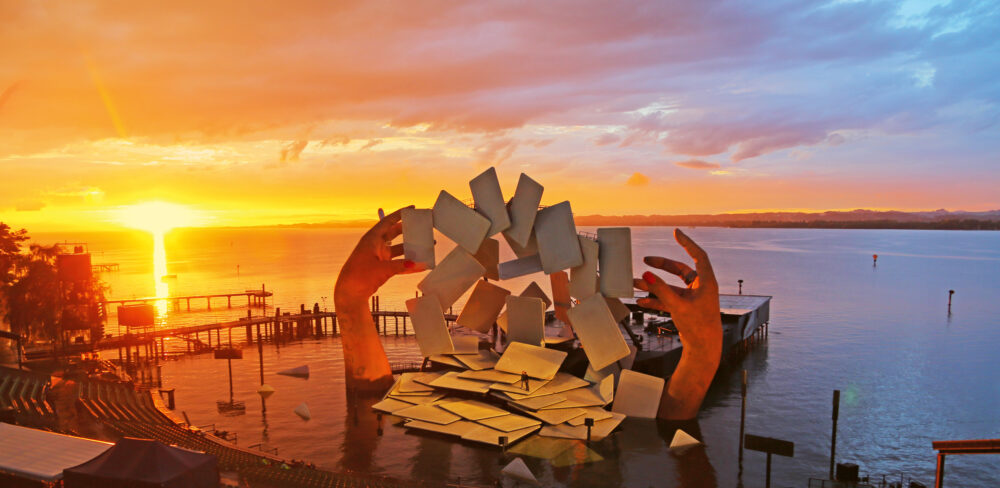 Large-scale outdoor installation of two large hands extending upwards from the ground, the left hand holds a cigarette. Several floating cards are suspended between them and more lay on the floor. The setting is at a pier by the water at sunset.
