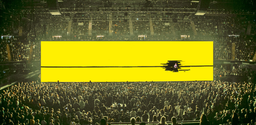 A large, indoor concert venue full of people with a large yellow rectangular screen in the center of the room; a black line cuts through the screen revealing a person towards the right.