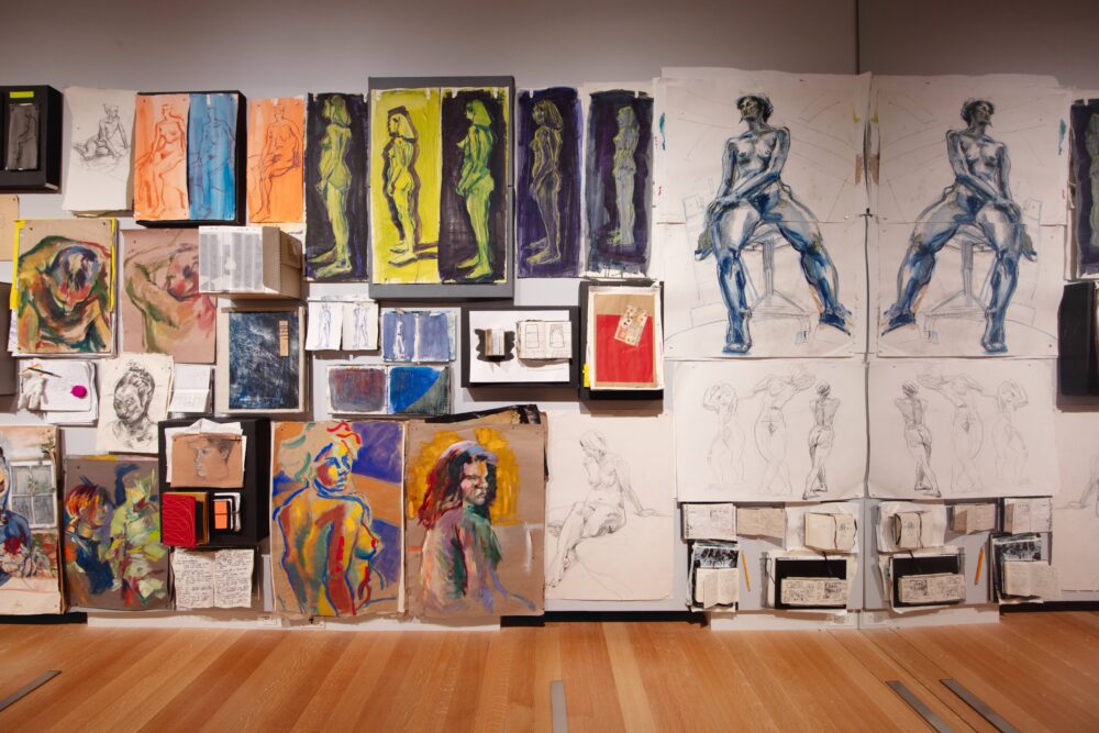 Straightforward view of a large-scale collage of artworks and sketchbooks pinned to a white wall. Some works are black and white whereas most are in color. Most of the imagery depicts figures, faces, and geometric forms.