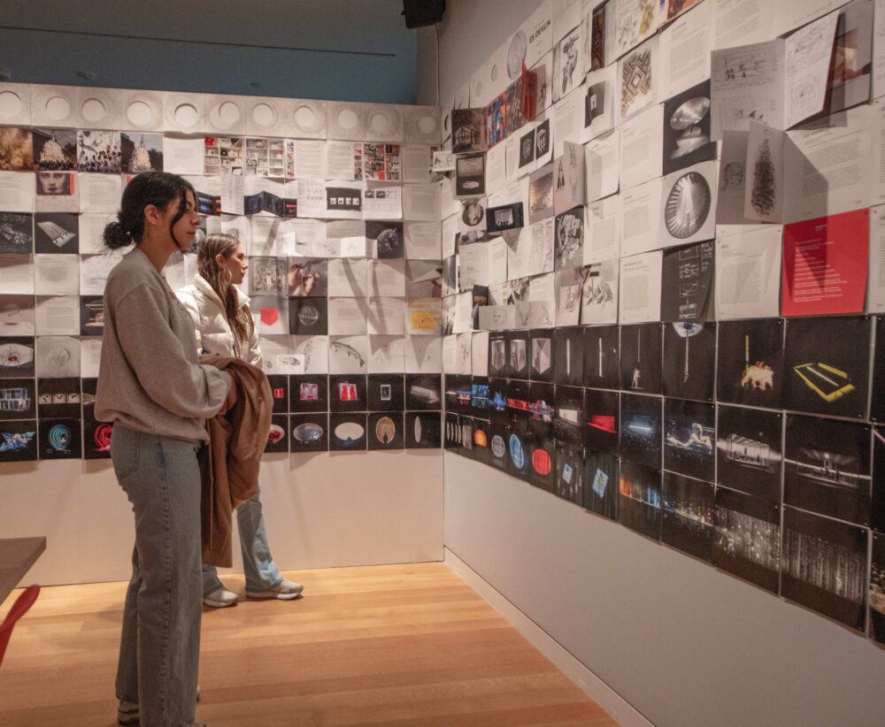 Two people are shown looking at two walls covered in a series of square pages with text and images that are lined up next to each other.