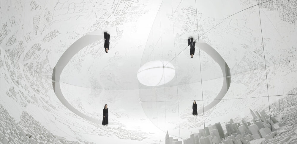 The illusion of a white spherical room is created with a mirrored ceiling and wall. A glowing white light centers the space surrounded by reflections of white models of a city and a person dressed in black, which is reflected four times.