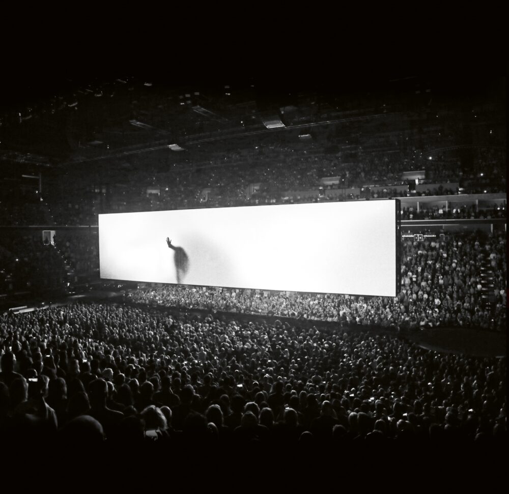 A large, indoor concert venue full of people with a large rectangular floating screen in the center of the room; the screen is mostly white with a large shadow raising its hand against the screen.