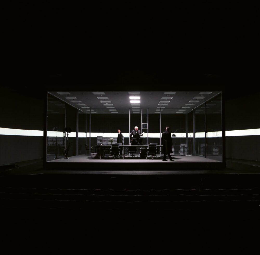 Black rectangular structure with no walls housing office furniture and a ceiling with several squares of which two are lit; inside are three people, one sitting on the table. Behind the structure is a stripe of white light that cuts across the black background.