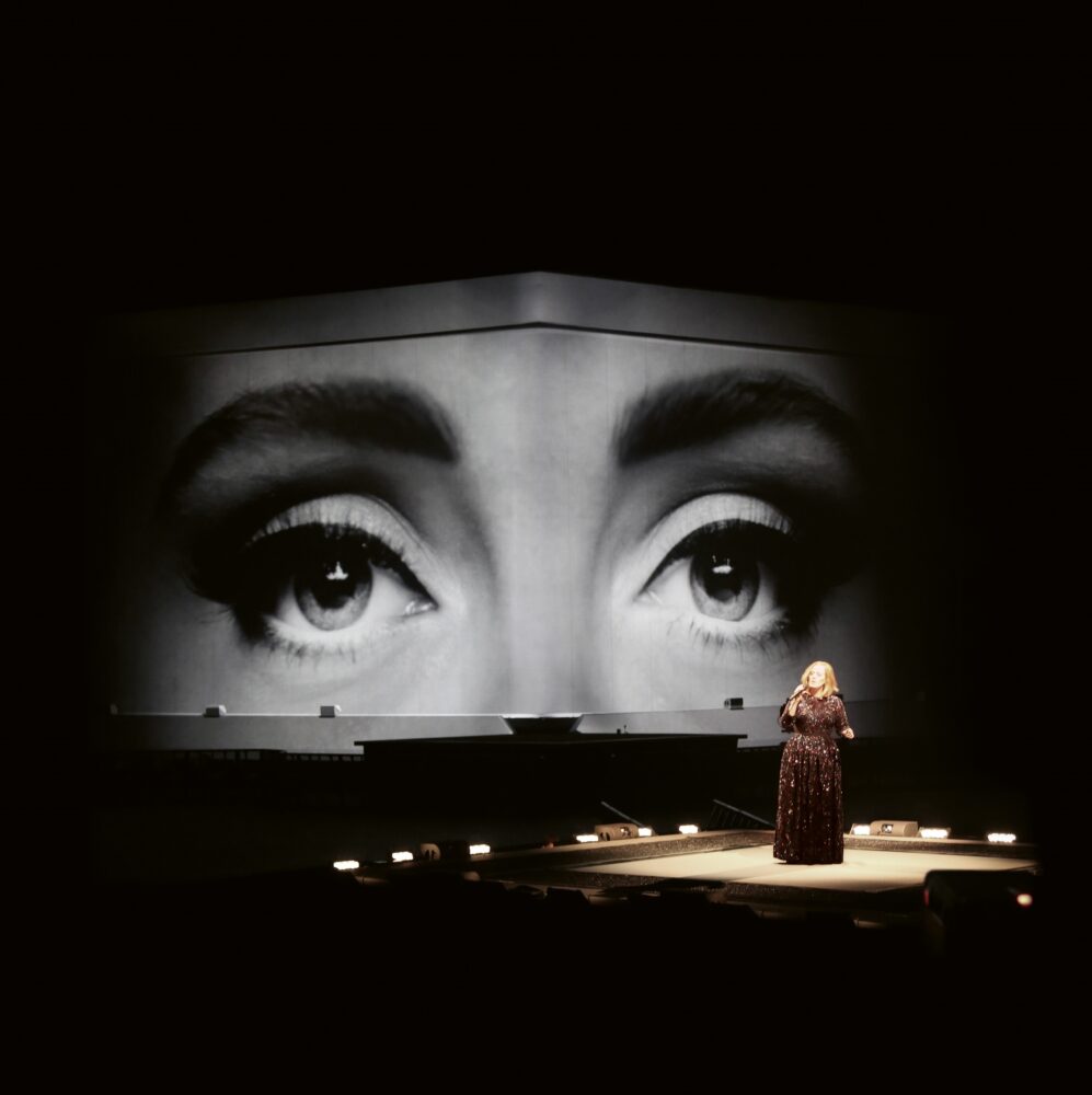 Singer Adele on stage in a dark room with a large, black-and-white projection of her eyes behind her.