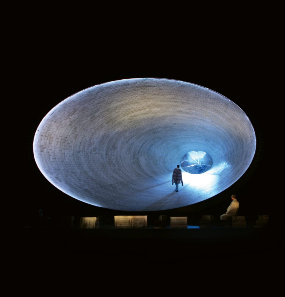 Large oval tunnel with gray, brick-textured surfaces above a black background. A person walks away from the viewer toward the opposite end of the tunnel which is illuminated.