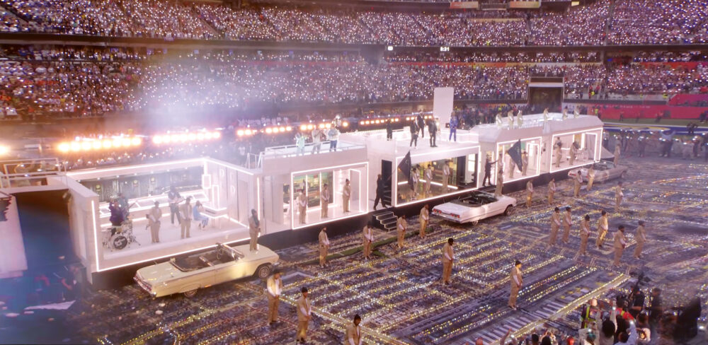 Large stadium venue full of people with a complex white rectangular structure in the center. The structure has open sides and outlines in white light. Several performers are dispersed throughout and three cars are parked next to the structure.