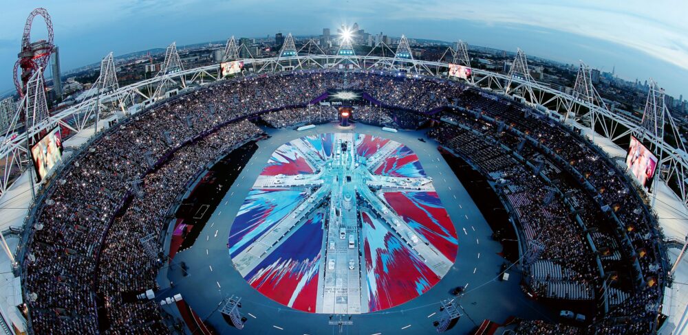 A large stadium full of people is seen from above. The center stage comprises a large oval with a red, blue, and white geometric design—an abstraction of the British flag.
