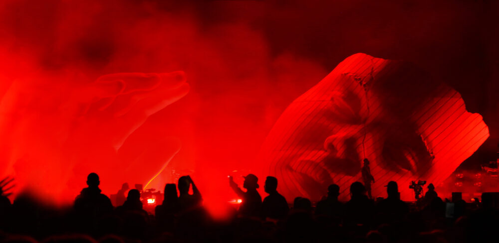 A large model of a hand and face are bathed in red light and surrounded by fog, foregrounded by a silhouette of a crowd of people.