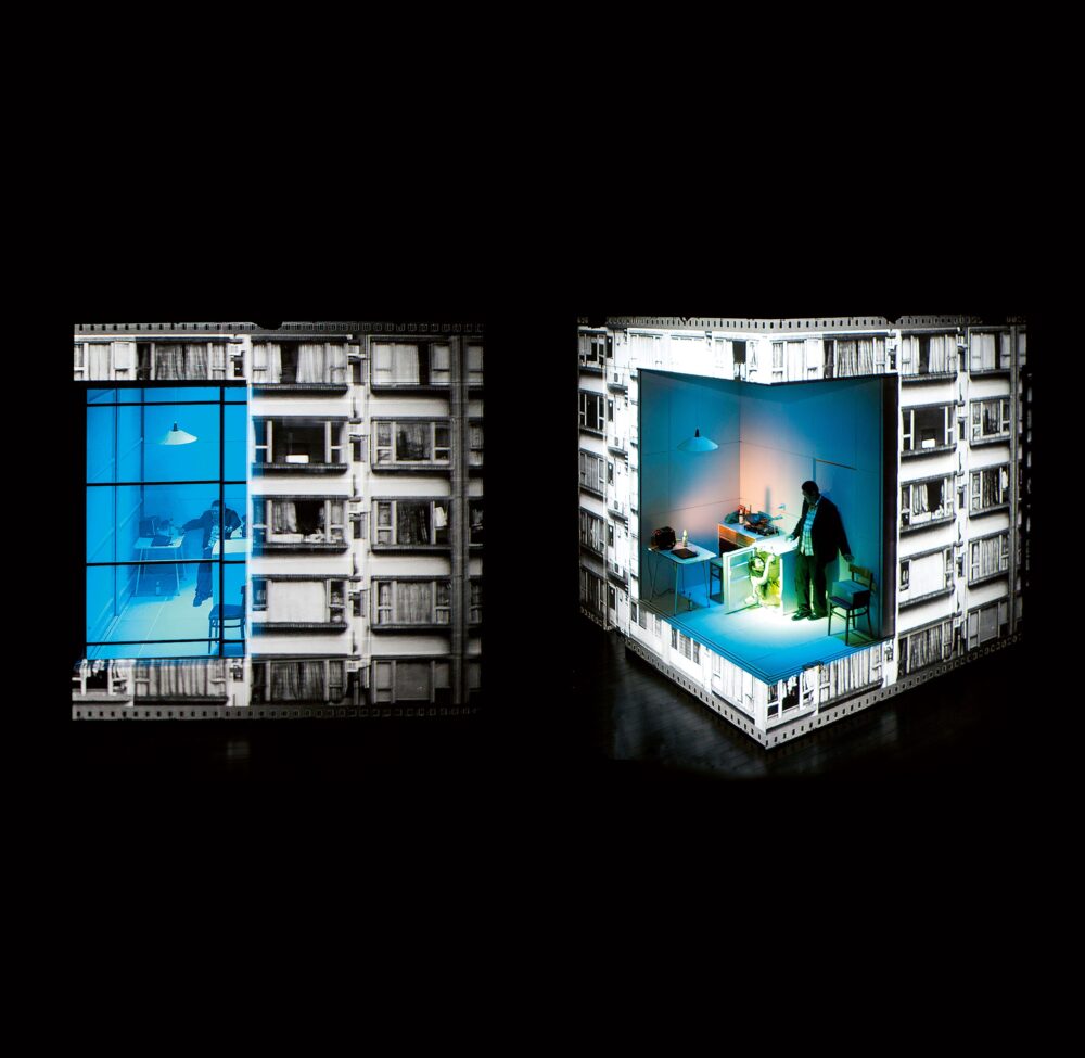 Two cubic structures shown from different angles: straight on and at an angle. Black-and-white windows are projected on the exteriors; both cubes have blue-lit interiors with a person standing inside.