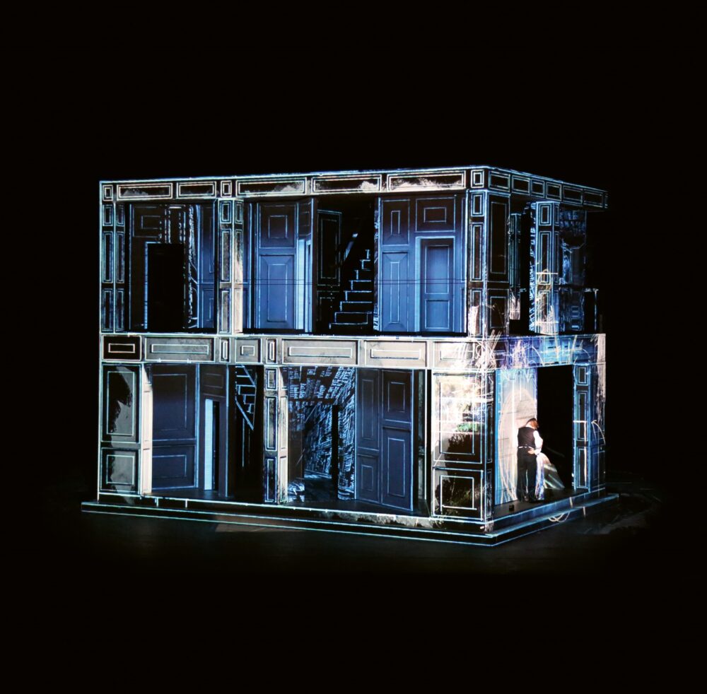 Rectangular structure with two levels sits in a black room. The structure is decorated with architectural elements like doors and windows that are projected onto the surfaces with white and blue light.