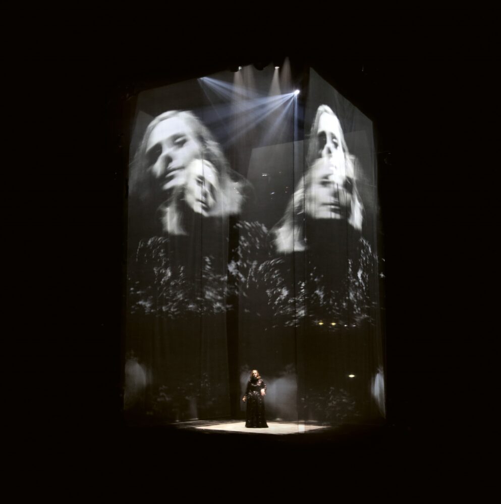 Singer Adele on stage in a dark room surrounded by four large-scale, black-and-white projections of her.