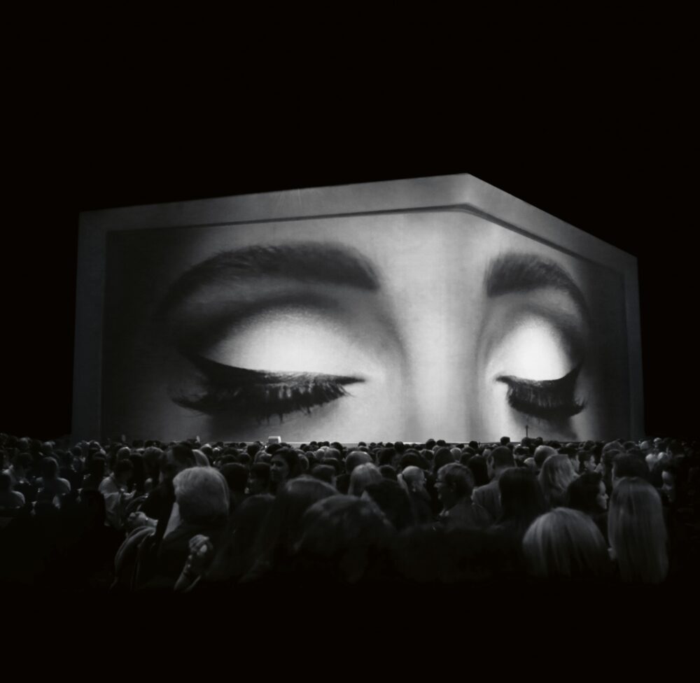 Black-and-white image of a large-scale projection of two closed eyes above a large crowd facing the screen.