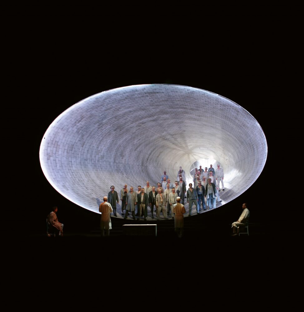 Large oval tunnel with gray, brick-textured surfaces above a black background. A crowd of people fills the tunnel and their proportional sizes indicate depth. Four people exist outside of the oval toward the bottom and are facing it.