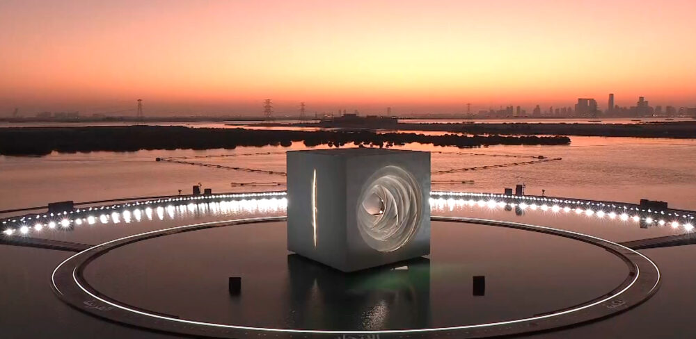 Large white cube, with different cutout designs on each side, is pictured outdoors on a smooth circular surface. The cube emits light and the floor has several other white lights. The background is a cityscape by the water at sunset.
