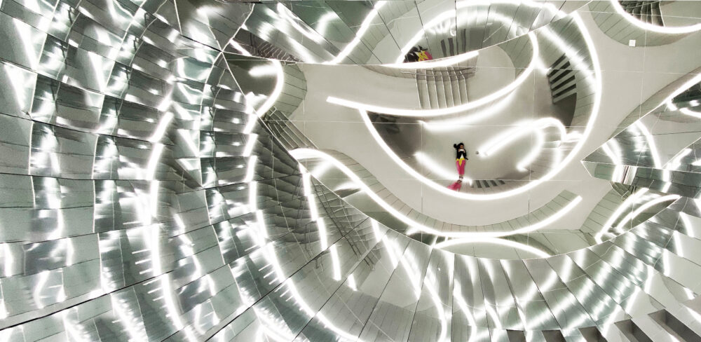 A series of reflective surfaces in a spiral, maze-like form; they are reflecting curved lines of white light.