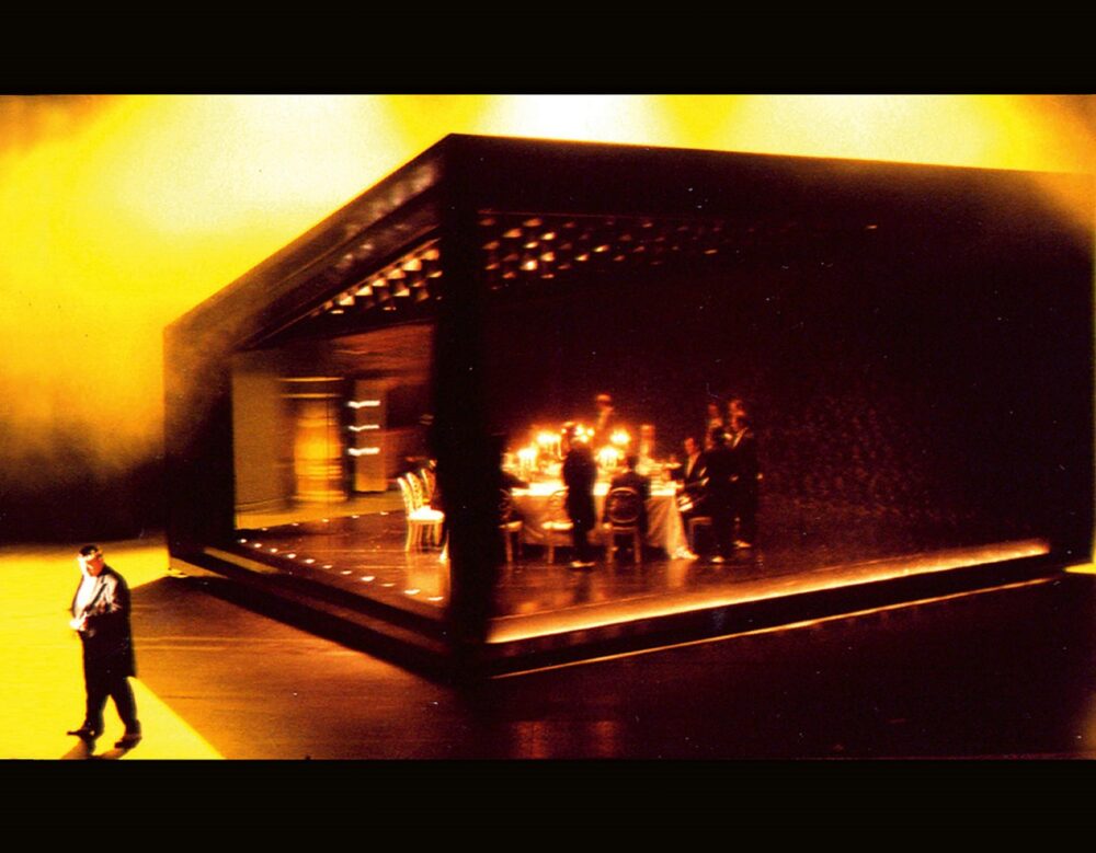 A large black trapezoidal structure houses a white dining table and chairs with a group of people dressed in suits. The structure sits on a flat surface and is surrounded by a yellow, hazy atmosphere. A person, closer to the viewer than the rest, is in the bottom left corner facing the viewer.