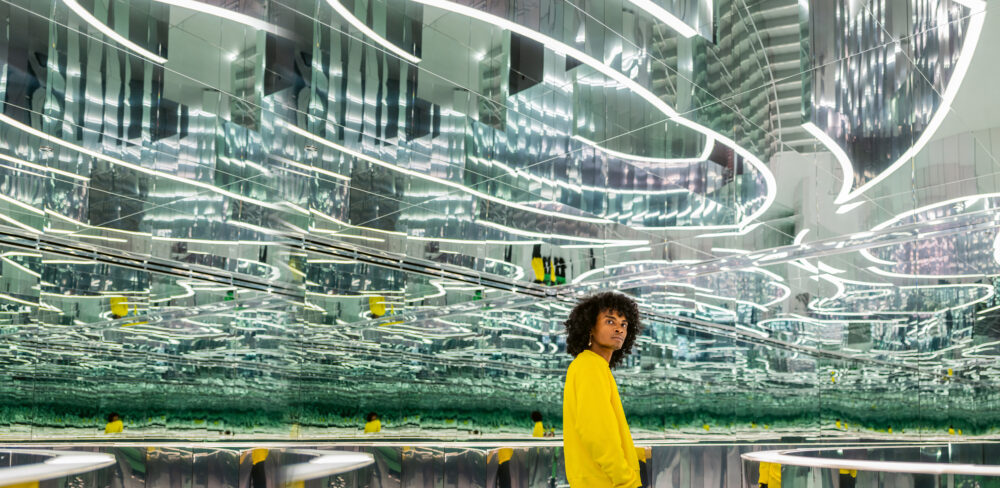 A person in a yellow long-sleeved shirt in a large room with a ceiling consisting of mirrors in a maze-like composition; the yellow shirt repeats throughout the space along with lines of white light.