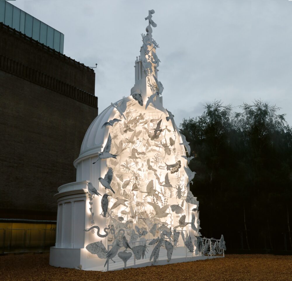 A large white architectural structure sits outdoors next to a building and trees. The installation is half of a dome-like form with white light glowing from inside that highlights countless silhouettes of flying birds, made of a similarly white material.