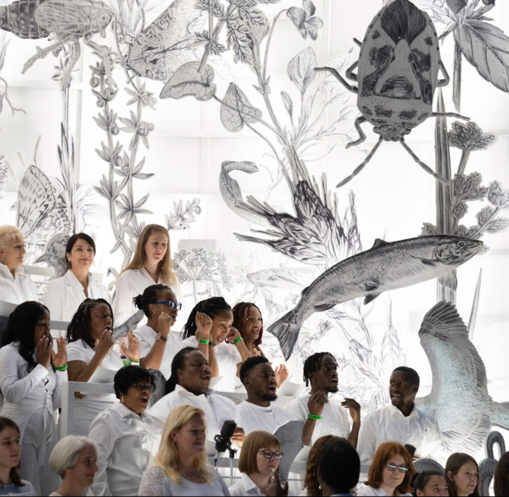Photograph of a group of people dressed in white against a background of realistic black and white drawings of plants and animals.