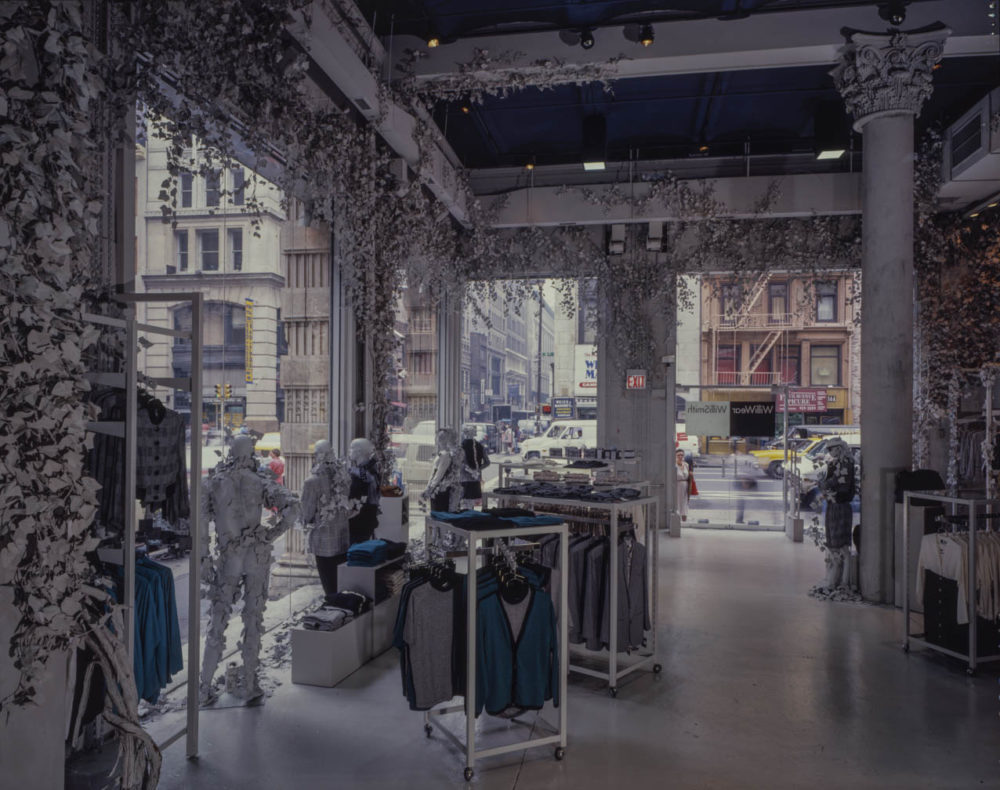 Interior of a gray clothing store with large display windows and gray ivy hanging from the walls and ceiling. Clothing racks and white stackable boxes display clothing. Multiple mannequins wear clothing with ivy leaves draped around them. At the front of the store is a black-and-white sign that reads, "WilliWear, Willi Smith."