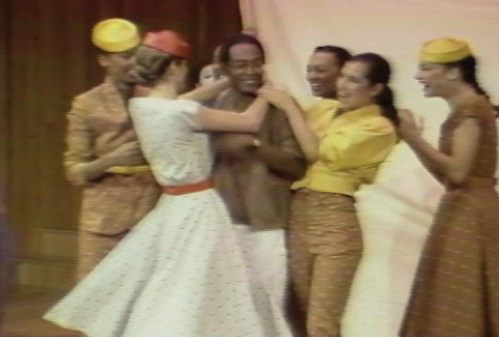 Six female models, and one mime, surround a smiling dark-skinned man. The models clap and smile in the man's direction, and two models hug the man.
