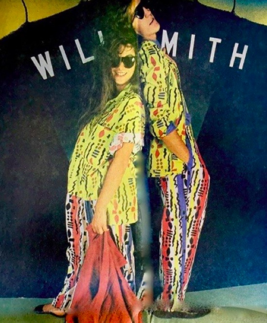 Photo of a woman and man leaning against each other, back-to-back, in colorful patterned clothing with text stating “Willi Smith” painted on a backdrop behind them.