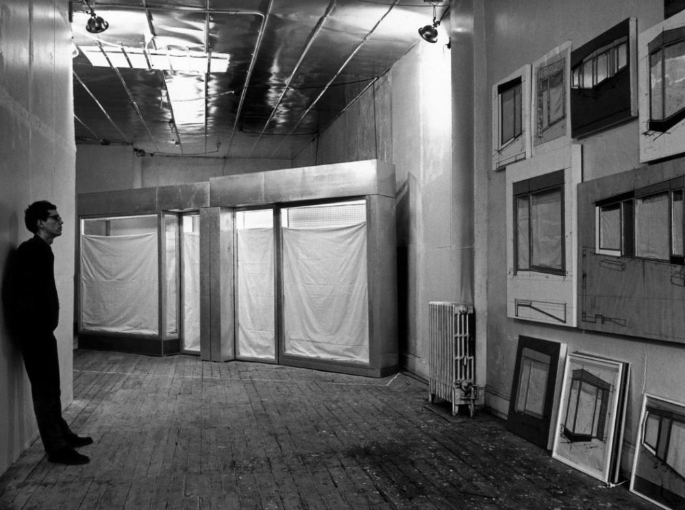 Black and white photo of man standing in a large industrial room against the wall on the left of the image. On the right wall are large sketches of a storefront. At the rear of the room is a full-scale mockup of the storefront.