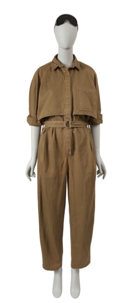 A khaki brown aviator style jumpsuit with a double ring belt. The jumpsuit features two overhanging panels on either side of the button placket that wrap around the waist to form a faux jacket from the side and back views. The mannequin is styled with a pair of black aviator goggles worn on top of the head