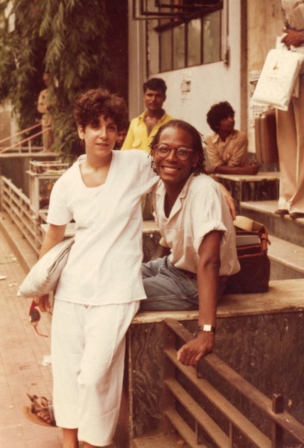 A light-skinned woman hugs a dark-skinned man. The woman wears a loose white shirt with white pants; the man wears round glasses, a button-up shirt with rolled sleeves, and grey pants. Two dark-skinned men in button-up shirts stand behind them in the background.