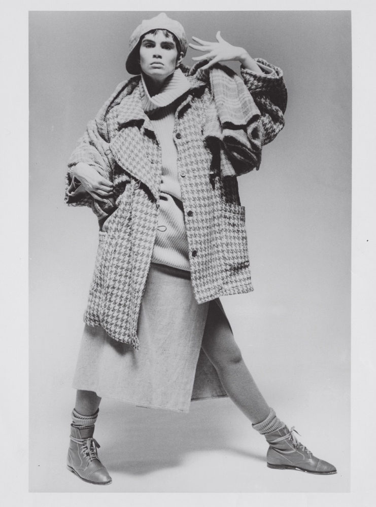 Grayscale photograph of model wearing houndstooth coat, thick turtleneck sweater, plaid scarf, skirt with side slit, and boots with thick socks
