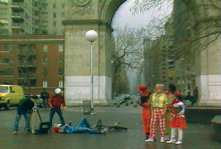 Still image from Made in New York film showing a staged bicycle incident in a park with an actor laying on the ground beside bike and other actors watching.