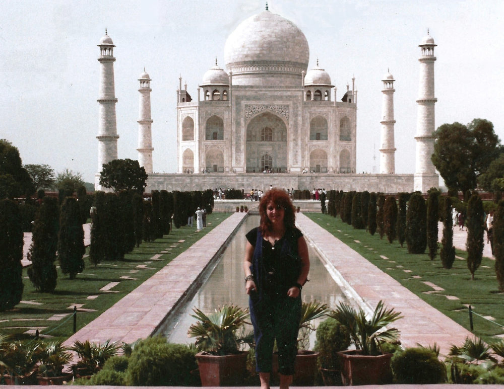 A light-skinned woman with red hair stands smiling in front of the Taj Mahal. The woman wears a black short-sleeve shirt with dark blue high-waisted pants and a dark blue vest.