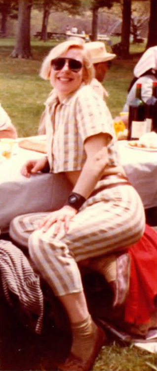 Photograph of a light-skinned woman with short blonde hair sitting at a long table in a park. The woman looks at the camera smiling and wears dark sunglasses, a striped short-sleeve crop top, and matching striped pants with brown flats.