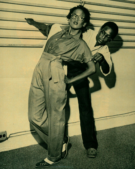 A yellow-toned photograph of two figures. In the foreground, a dark-skinned model poses and looks off to the right. Behind her, a dark-skinned man adjusts her clothing.
