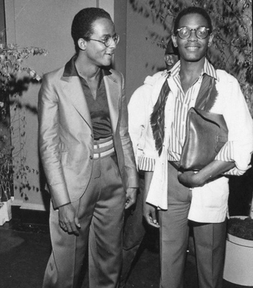 Grayscale photograph of two Black men standing together, smiling. Both men wear glasses; the man on the left wears a shimmering suit with a button-up shirt; the man on the right wears a striped button-up shirt with a white jacket and dress pants.