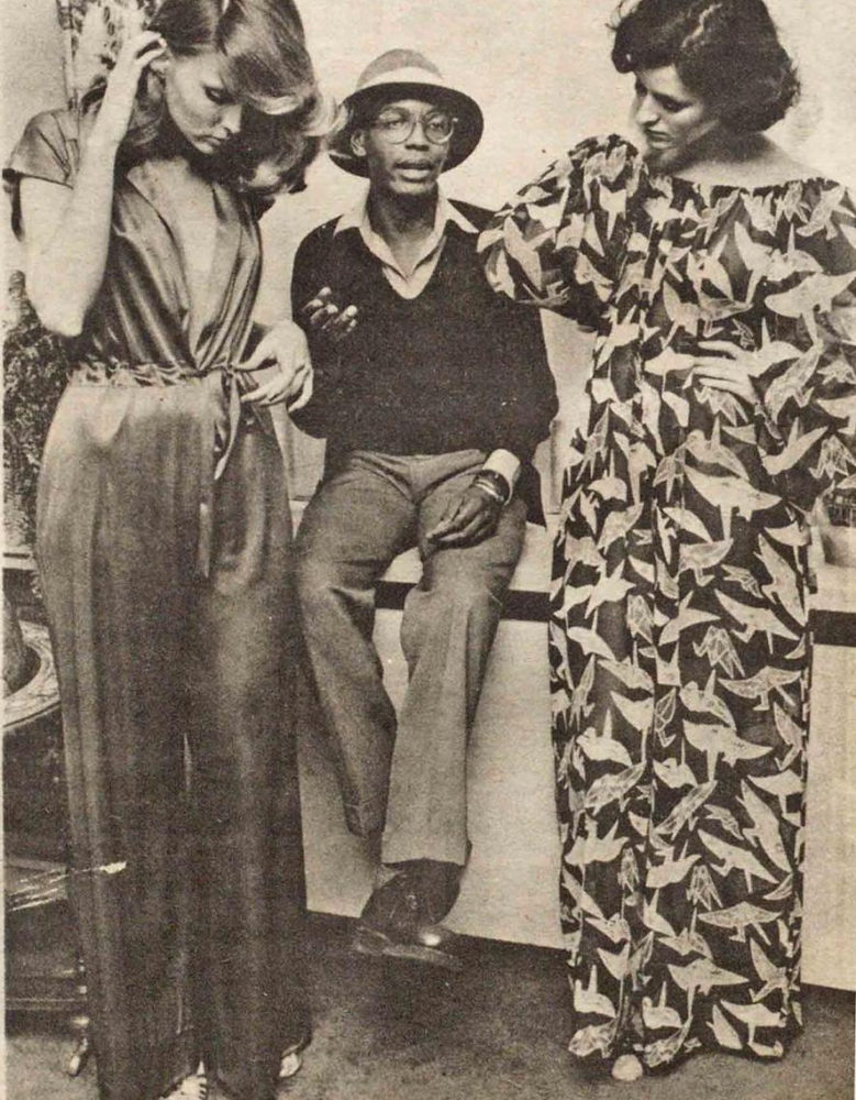Black and white image of three figures. In the center, a dark-skinned man sits on a counter, speaking. On his right, a woman stands in a long dark jumpsuit. On his left, a woman stands in a dress with paper crane patterns.