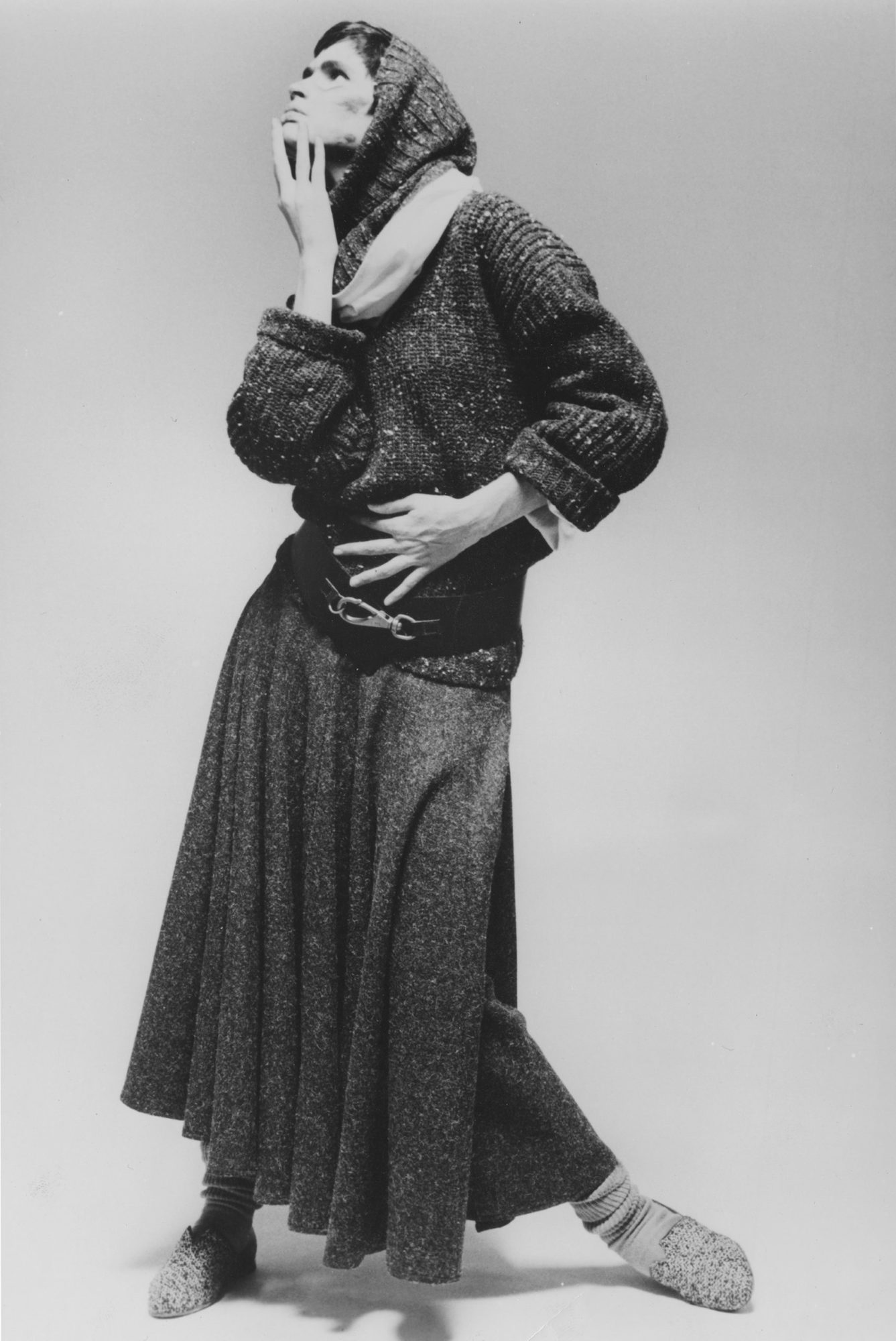 Grayscale image of a model posing dramatically wearing a dark hooded sweater, long wool skirt, large decorative belt, thick socks, and flat shoes