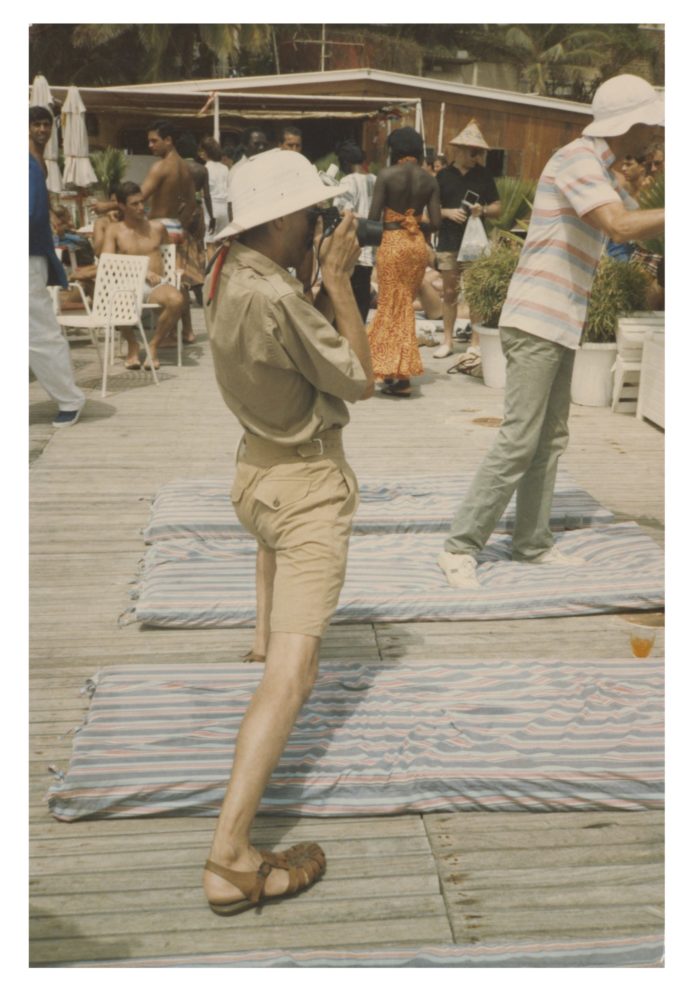 A man taking a photograph wearing a pith helmet, tan shirt with rolled up sleeves, and khaki shorts
